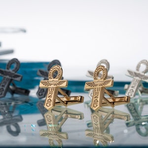 May include: Four gold cufflinks with an ankh design, two are on a blue glass surface and two are reflected in the glass.