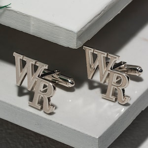 May include: Two silver cufflinks, each displaying the letters "WR" in a bold, serif font. The cufflinks are positioned on a white, textured surface, with a minimalist aesthetic. The design is simple and elegant, suitable for formal wear.
