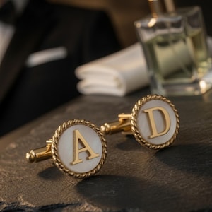 Op de afbeelding: Gouden manchetknopen met de letters "A" en "D" op een witte achtergrond, omlijst door een touwachtig ontwerp. De manchetknopen worden gepresenteerd op een donkere ondergrond, met de tekst "Custom Made For You" eronder.