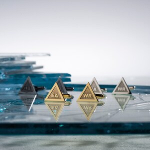 May include: Gold and silver triangular cufflinks with engraved initials. The cufflinks are arranged on a blue glass surface.