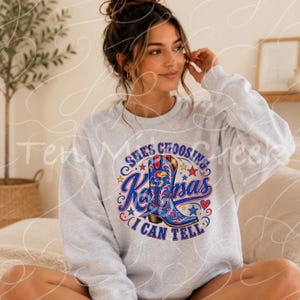 Puede incluir: Una sudadera gris claro de cuello redondo con un estampado gráfico de una bota vaquera azul con detalles en rojo y blanco. El texto "SHE'S CHOOSING KANSAS I CAN TELL" aparece en letras azules y rojas, rodeado de pequeñas estrellas y corazones.