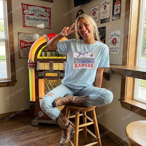Puede incluir: Una camiseta azul claro con un estampado gráfico de un estadio y el texto "They're coming" y "KANSAS I CAN TELL" en rojo y azul, combinada con pantalones vaqueros azul claro y botas vaqueras de cuero marrón con diseño de punta.