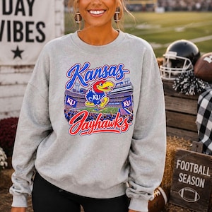 May include: A light grey crewneck sweatshirt featuring a graphic print of a stadium with the Kansas Jayhawks mascot in the centre. The text reads Kansas in blue script and Jayhawks in red script, with blue KU flags visible in the stadium stands.