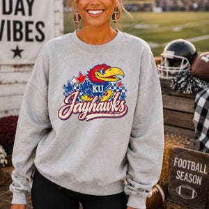 May include: A light grey crewneck sweatshirt featuring a distressed graphic of a blue and red Jayhawk mascot with the text KU and Jayhawks in a stylised font, surrounded by blue and red stars and a chequered pattern on the front chest area.