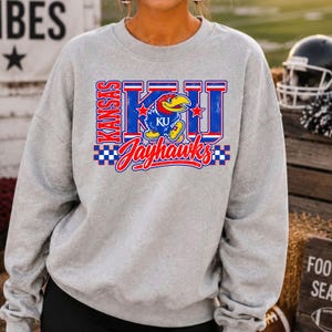 May include: A light grey crewneck sweatshirt featuring a large graphic on the chest. The design includes the text Kansas, KU, and Jayhawks in red, blue, and white, along with a blue Jayhawk mascot illustration and red star accents.