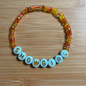 Puede incluir: Una pulsera de cuentas con la palabra "SHOWGIRL" escrita en letras doradas sobre cuentas celeste claro. La pulsera est&aacute; hecha de peque&ntilde;as cuentas coloridas en tonos naranja, amarillo y rosa.