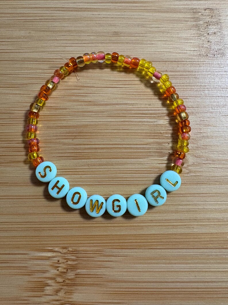 Puede incluir: Una pulsera de cuentas con la palabra "SHOWGIRL" escrita en letras doradas sobre cuentas celeste claro. La pulsera est&aacute; hecha de peque&ntilde;as cuentas coloridas en tonos naranja, amarillo y rosa.