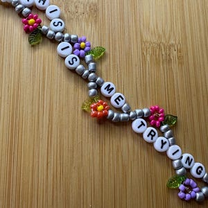 May include: A beaded bracelet spelling out "THIS IS ME TRYING" with white letter beads. The bracelet has silver beads and colourful flower accents in pink, purple, and yellow.
