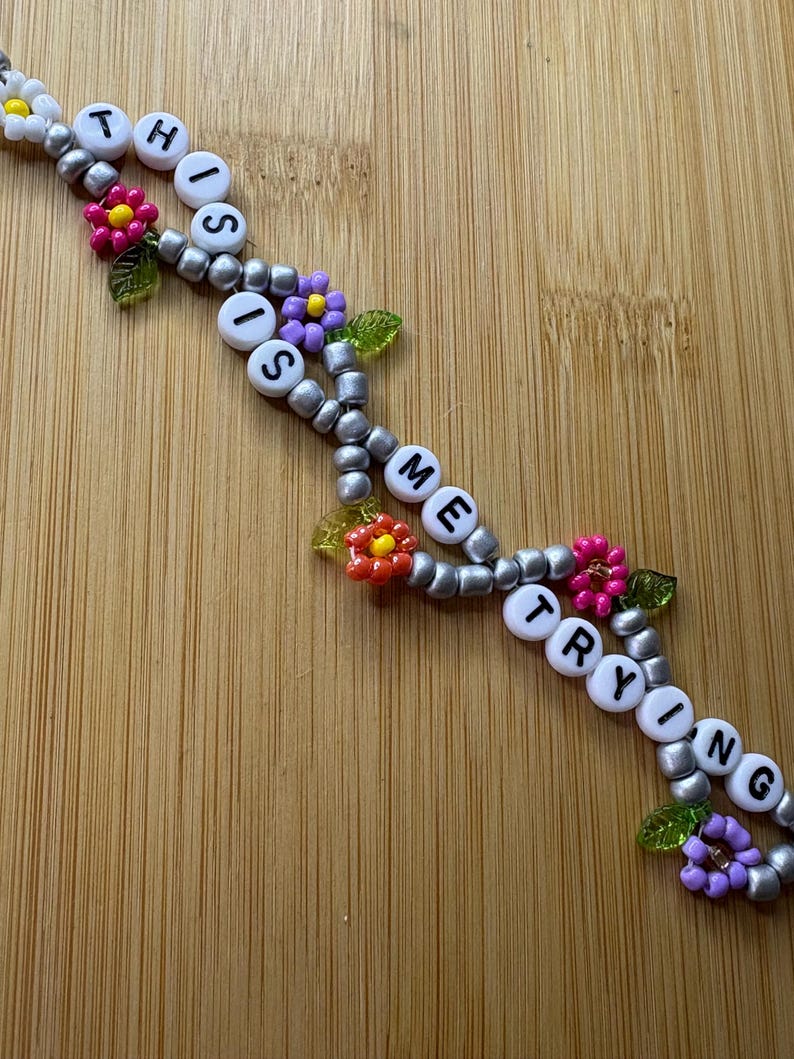 May include: A beaded bracelet spelling out "THIS IS ME TRYING" with white letter beads. The bracelet has silver beads and colourful flower accents in pink, purple, and yellow.