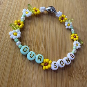 May include: A beaded bracelet with the words "OUR SONG" spelled out in white and light blue beads. The bracelet features yellow and white flower beads, green leaf beads, and a silver magnetic clasp.