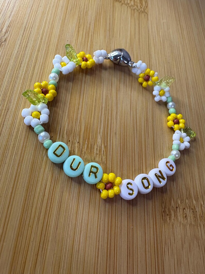 May include: A beaded bracelet with the words "OUR SONG" spelled out in white and light blue beads. The bracelet features yellow and white flower beads, green leaf beads, and a silver magnetic clasp.
