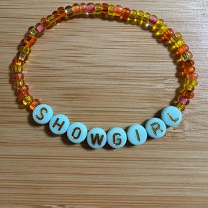 Puede incluir: Una pulsera de cuentas con la palabra "SHOWGIRL" escrita en letras doradas sobre cuentas celestes. La pulsera presenta un degradado de cuentas naranjas, amarillas y rosas.