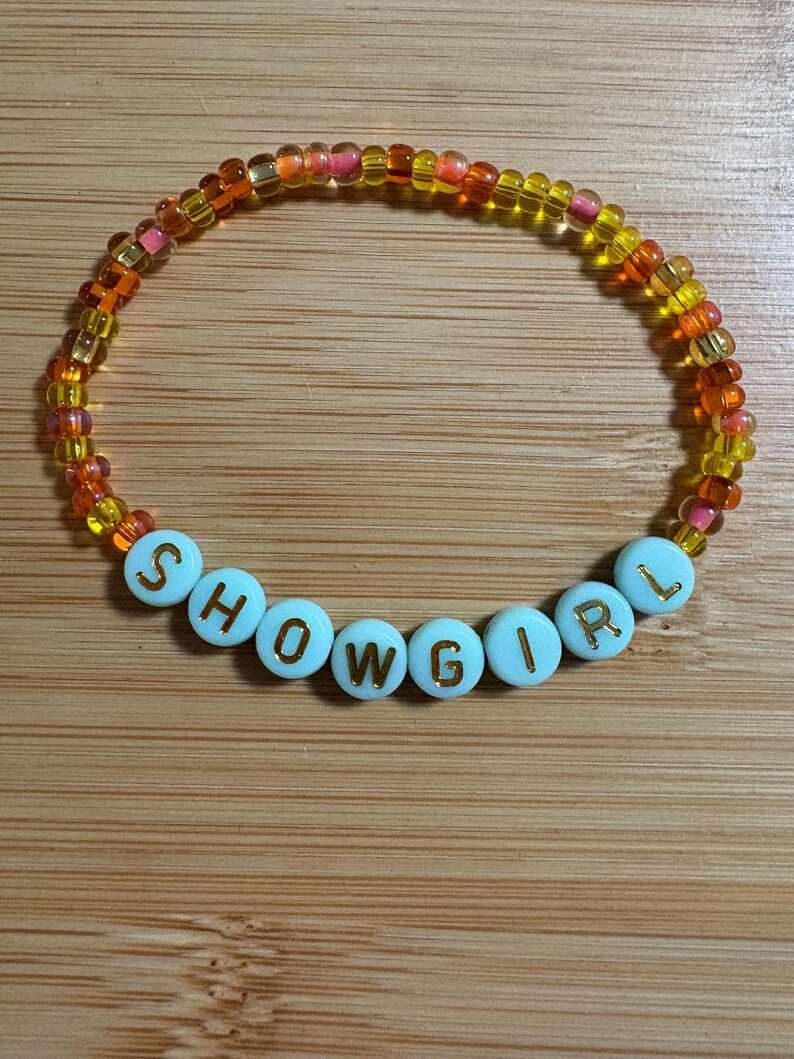Puede incluir: Una pulsera de cuentas con la palabra "SHOWGIRL" escrita en letras doradas sobre cuentas celestes. La pulsera presenta un degradado de cuentas naranjas, amarillas y rosas.
