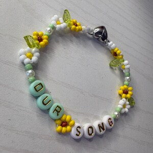 May include: A beaded bracelet with the words "OUR SONG" spelled out in white and mint green beads. The bracelet features yellow and white flower accents, green leaf beads, and a silver heart-shaped clasp.
