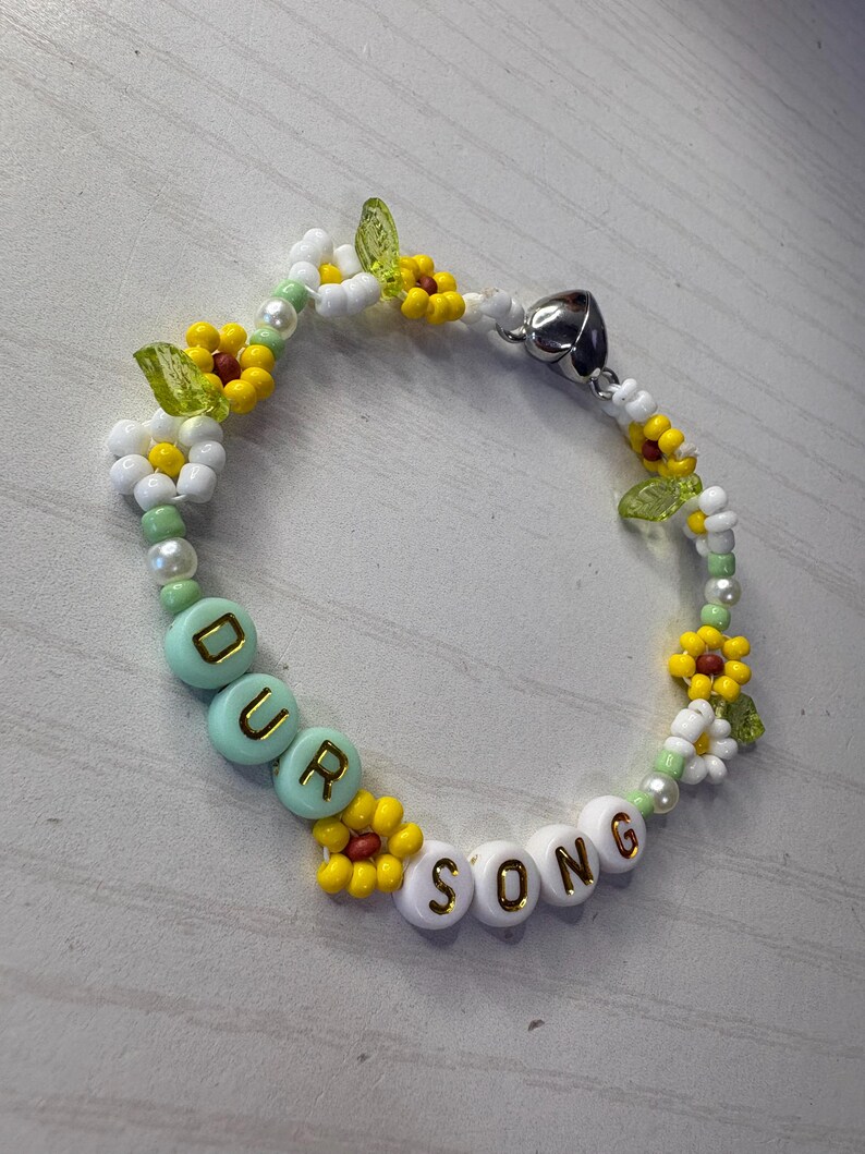 May include: A beaded bracelet with the words "OUR SONG" spelled out in white and mint green beads. The bracelet features yellow and white flower accents, green leaf beads, and a silver heart-shaped clasp.