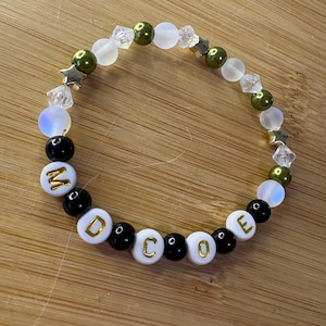 May include: A beaded bracelet with the word "MODE" spelled out in gold letters on white beads. The bracelet features black, white, iridescent, and olive green beads, along with star-shaped and crystal accents.