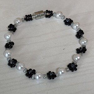 May include: A beaded bracelet featuring white faux pearls, black flower-shaped beads, and small gray beads. The bracelet has a magnetic clasp and is designed for a delicate, feminine look.