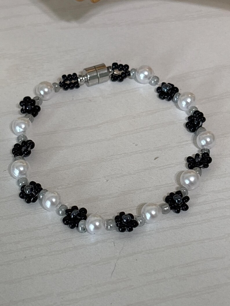 May include: A beaded bracelet featuring white faux pearls, black flower-shaped beads, and small gray beads. The bracelet has a magnetic clasp and is designed for a delicate, feminine look.