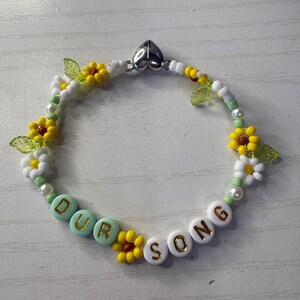 May include: A beaded bracelet with the words "OUR SONG" spelled out in white and turquoise beads. The bracelet features yellow and white flower accents, green leaf beads, and a silver heart-shaped clasp.
