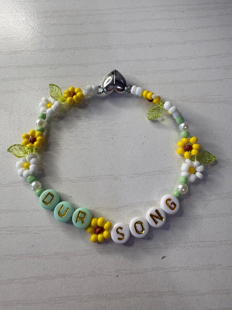 May include: A beaded bracelet with the words "OUR SONG" spelled out in white and turquoise beads. The bracelet features yellow and white flower accents, green leaf beads, and a silver heart-shaped clasp.