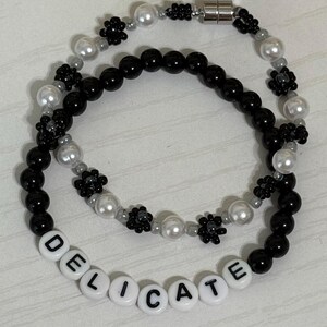 May include: Two beaded bracelets. One bracelet features black and white beads, with the word "DELICATE" spelled out in white letter beads. The other bracelet has black beads, white pearl beads, and small black flower accents. The bracelets are connected with a magnetic clasp.