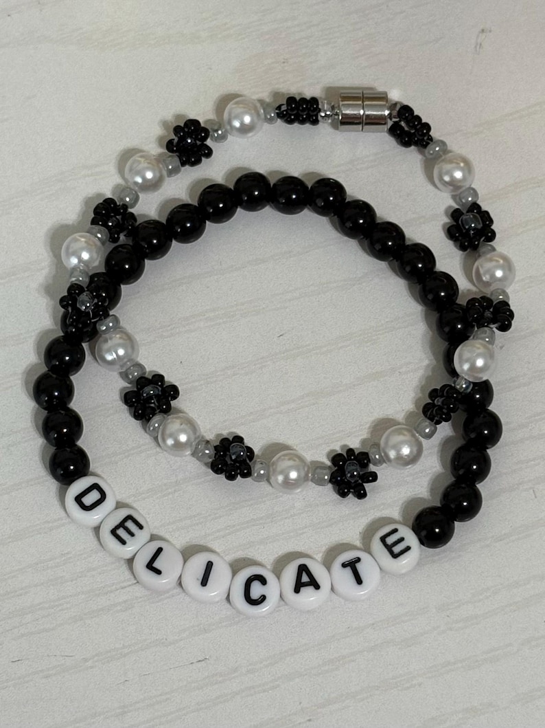 May include: Two beaded bracelets. One bracelet features black and white beads, with the word "DELICATE" spelled out in white letter beads. The other bracelet has black beads, white pearl beads, and small black flower accents. The bracelets are connected with a magnetic clasp.
