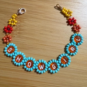 May include: A beaded bracelet with the word "SHOWGIRL". The bracelet features turquoise flower-shaped beads with orange centers and white letter beads. Additional beads are yellow, red, and orange.