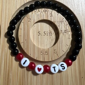 Op de afbeelding: Zwart kralenarmband met rode accentkralen en witte letterkralen met de tekst "I ❤️ TS". De armband wordt getoond op een houten meetinstrument met een cirkel met een diameter van 14 cm.