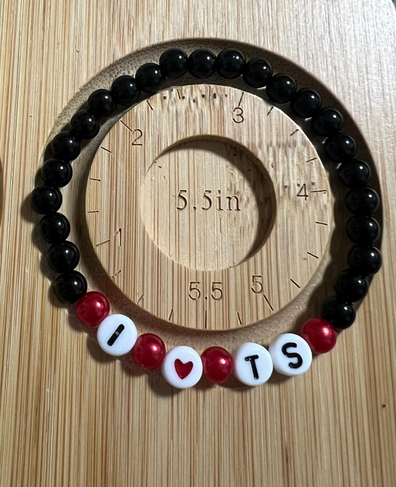 Op de afbeelding: Zwart kralenarmband met rode accentkralen en witte letterkralen met de tekst "I ❤️ TS". De armband wordt getoond op een houten meetinstrument met een cirkel met een diameter van 14 cm.