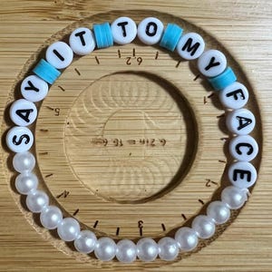 May include: A white beaded bracelet with blue and white letter beads spelling out "Say It To My Face".