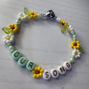 May include: A beaded bracelet with the words "OUR SONG" spelled out in white and light blue beads. The bracelet features yellow and white flower beads, green leaf beads, and a silver heart-shaped clasp.