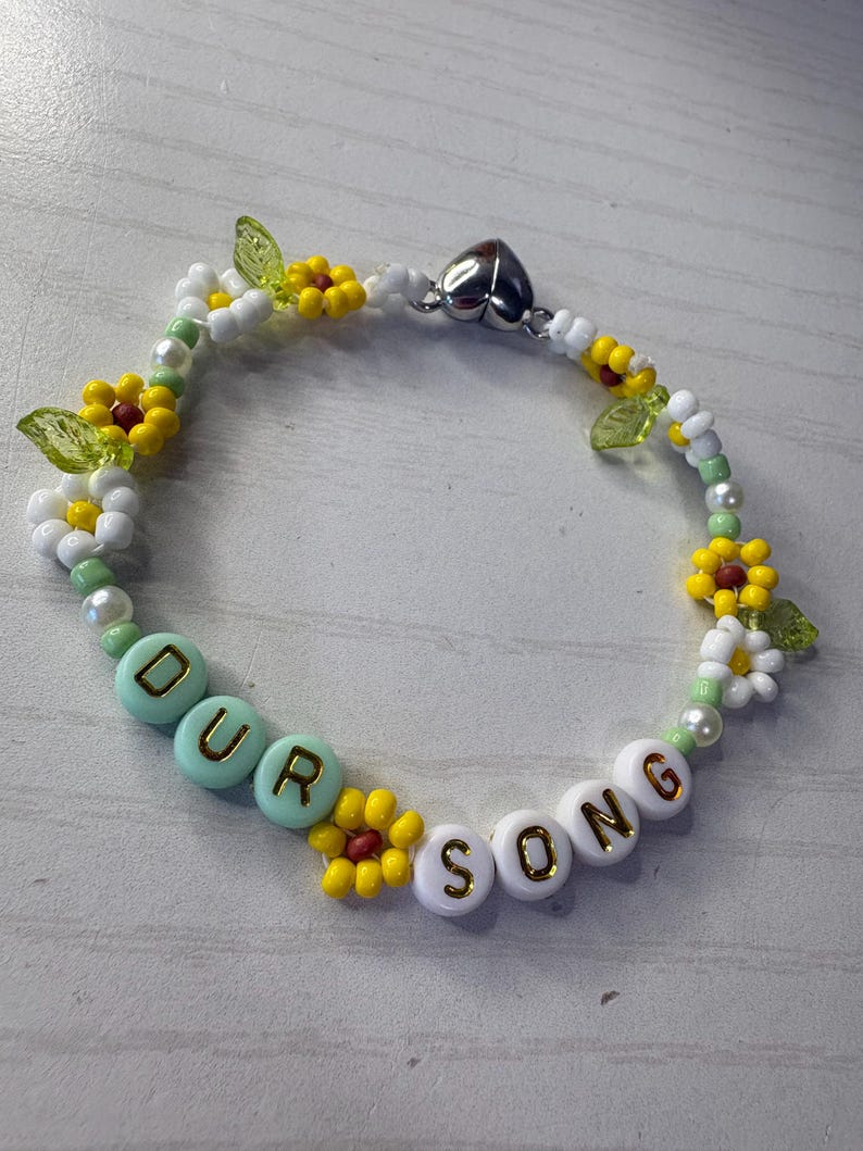 May include: A beaded bracelet with the words "OUR SONG" spelled out in white and light blue beads. The bracelet features yellow and white flower beads, green leaf beads, and a silver heart-shaped clasp.