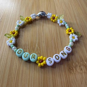 May include: A beaded bracelet with the words "OUR SONG" spelled out in white and mint green letter beads. The bracelet features yellow and white flower beads, green leaf beads, and a silver magnetic clasp.