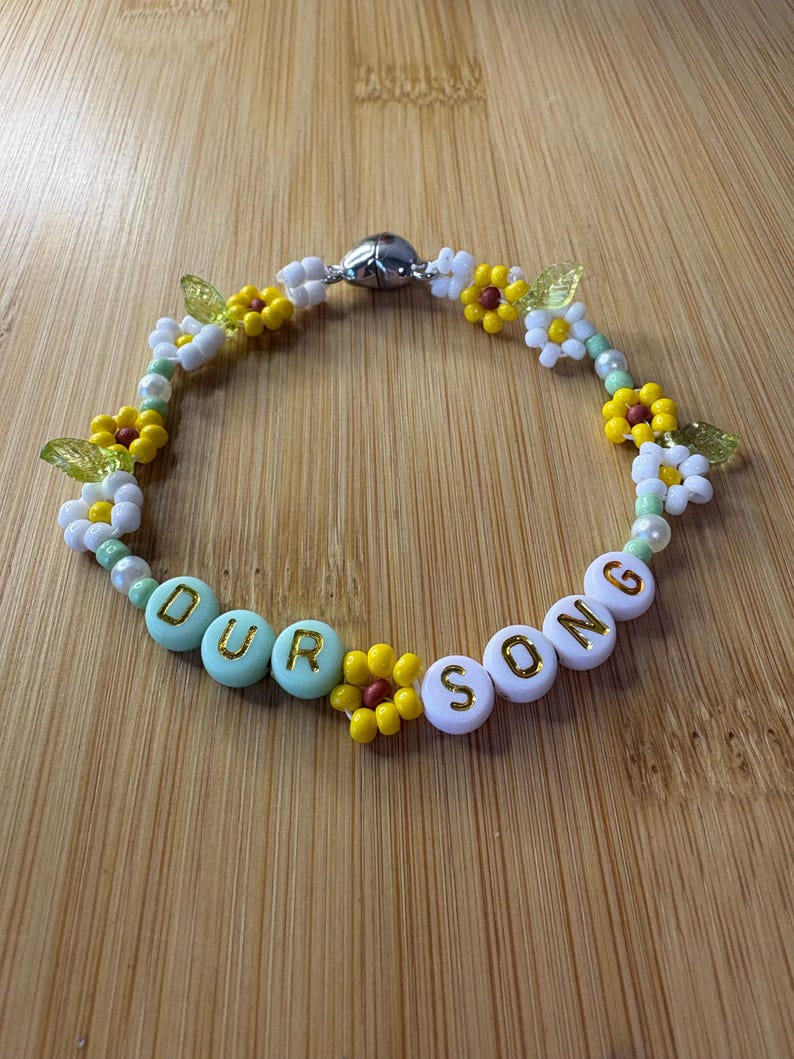 May include: A beaded bracelet with the words "OUR SONG" spelled out in white and mint green letter beads. The bracelet features yellow and white flower beads, green leaf beads, and a silver magnetic clasp.