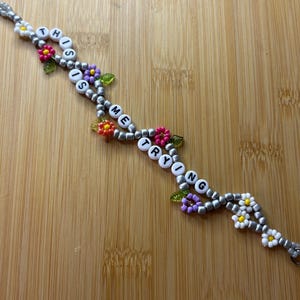 May include: A beaded bracelet spelling "THIS IS ME TRYING" with white letter beads. The bracelet has silver beads and is decorated with colourful flower and leaf accents.