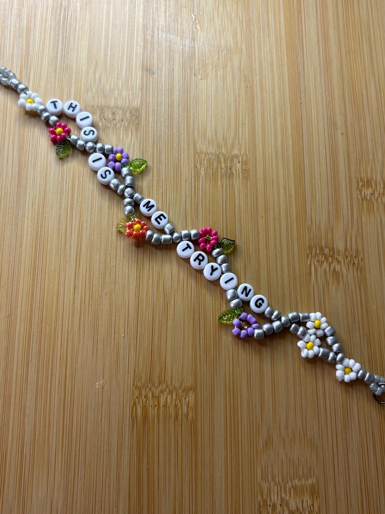 May include: A beaded bracelet spelling "THIS IS ME TRYING" with white letter beads. The bracelet has silver beads and is decorated with colourful flower and leaf accents.