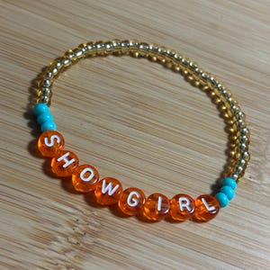 May include: A beaded bracelet with the word "SHOWGIRL" spelled out in orange letter beads. The bracelet features gold and turquoise beads, strung on a clear elastic band. The bracelet is displayed on a wooden surface.