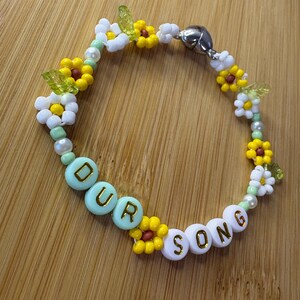 May include: A beaded bracelet with the words "OUR SONG" spelled out in white and light blue beads. The bracelet features yellow and white flower beads, green leaf beads, and a silver magnetic clasp.