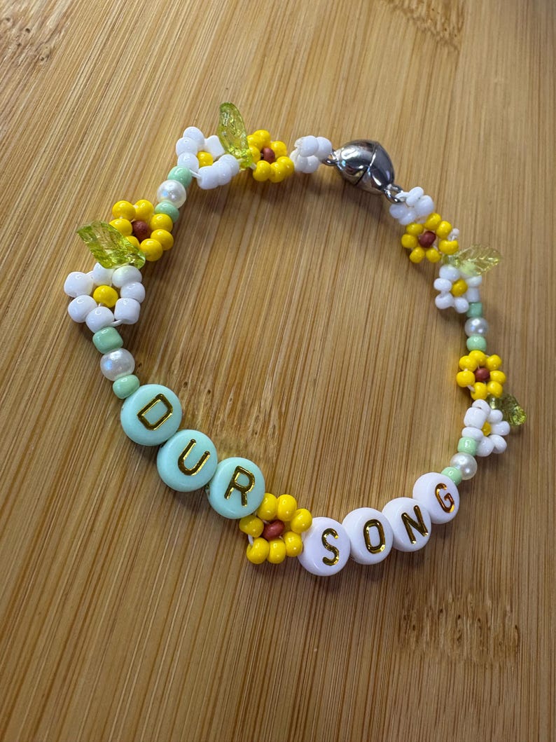 May include: A beaded bracelet with the words "OUR SONG" spelled out in white and light blue beads. The bracelet features yellow and white flower beads, green leaf beads, and a silver magnetic clasp.