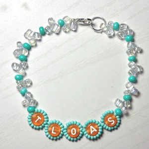 May include: A beaded bracelet with clear, turquoise, and orange beads. The bracelet features the letters "TLOAS" in orange circles. The bracelet is secured with a silver clasp. The beads are arranged in a floral pattern.