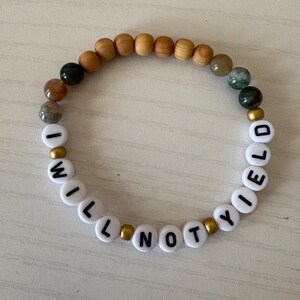 May include: A beaded bracelet with the words "I WILL NOT YIELD" spelled out in white letter beads. The bracelet also features wooden and gemstone beads, and gold-colored spacers. The bracelet is on a light-colored surface.