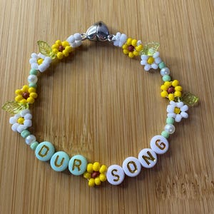 May include: A beaded bracelet with the words "OUR SONG" spelled out in white and turquoise beads. The bracelet is decorated with yellow and white flower beads, green leaf beads, and small white beads. The clasp is silver.