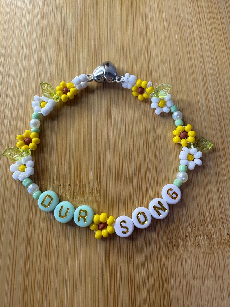 May include: A beaded bracelet with the words "OUR SONG" spelled out in white and turquoise beads. The bracelet is decorated with yellow and white flower beads, green leaf beads, and small white beads. The clasp is silver.