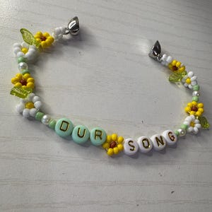 May include: A beaded bracelet with the words "OUR SONG" spelled out in gold letters. The bracelet features yellow and white flower beads, green leaf beads, and white beads. The bracelet has silver clasps.