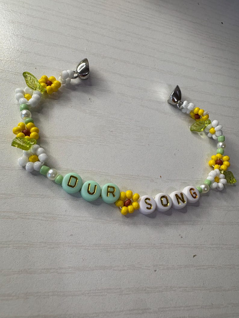 May include: A beaded bracelet with the words "OUR SONG" spelled out in gold letters. The bracelet features yellow and white flower beads, green leaf beads, and white beads. The bracelet has silver clasps.