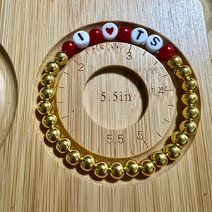 Op de afbeelding: Een goudkleurige kralenarmband met de letters "I", een hart, "T" en "S" gespeld in witte letterkralen en rode kralen. De armband heeft een diameter van ongeveer 14 cm. De armband ligt op een houten oppervlak.