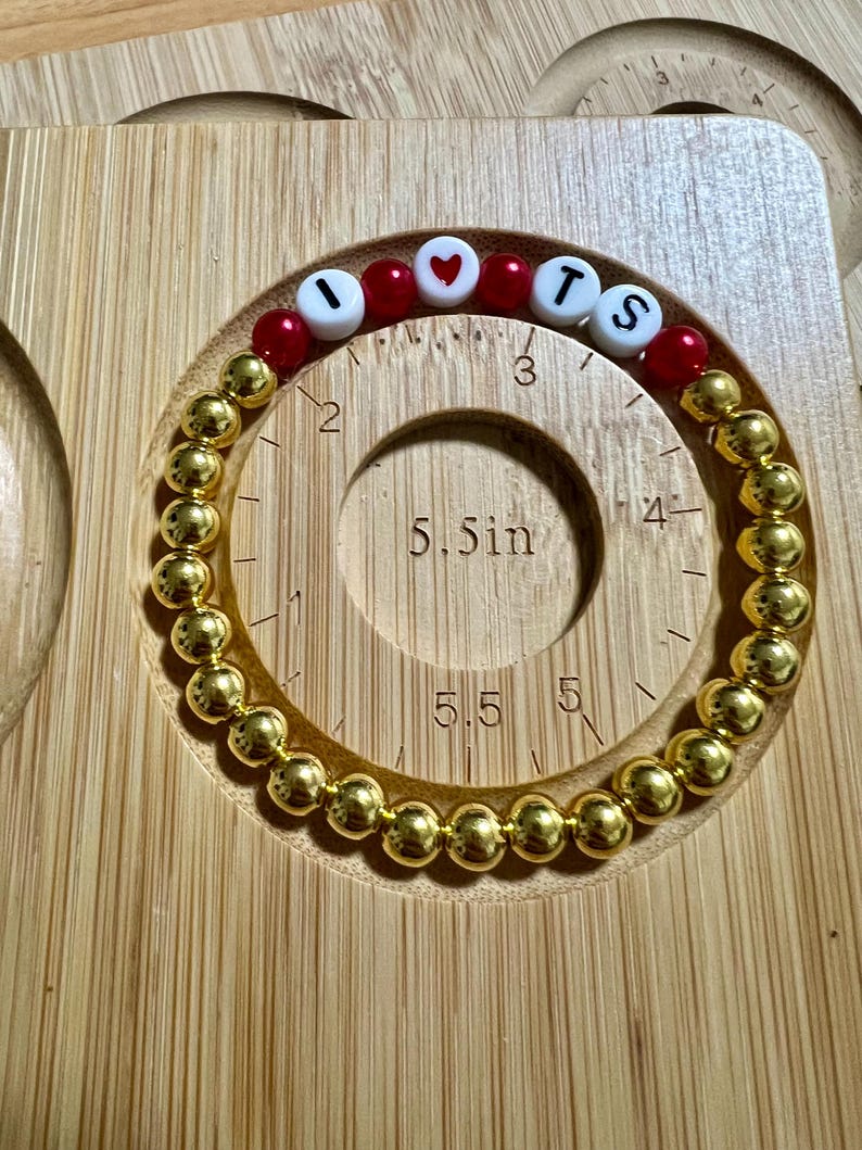 Op de afbeelding: Een goudkleurige kralenarmband met de letters "I", een hart, "T" en "S" gespeld in witte letterkralen en rode kralen. De armband heeft een diameter van ongeveer 14 cm. De armband ligt op een houten oppervlak.