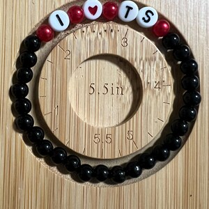 Op de afbeelding: Een zwart kralenarmband met de letters "I ❤️ TS" gespeld in witte letterkralen en rode hartkralen. De armband wordt weergegeven op een houten meetinstrument met een diameter van 14 cm.