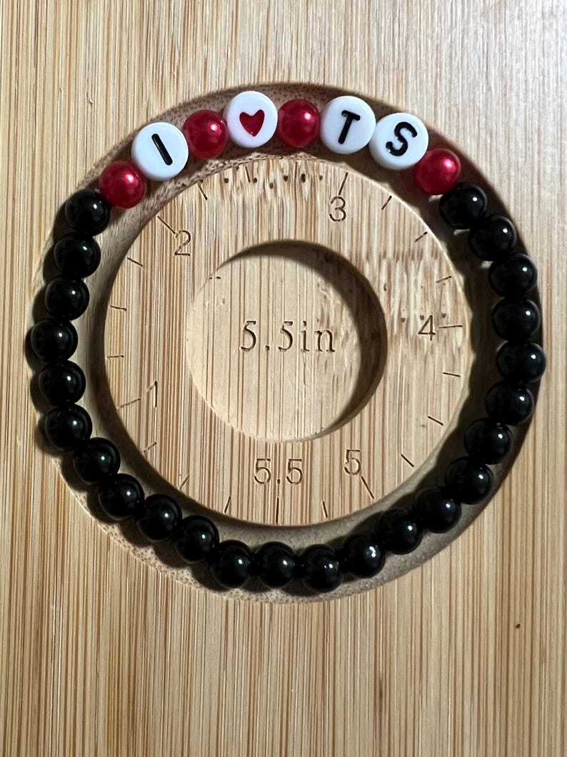 Op de afbeelding: Een zwart kralenarmband met de letters "I ❤️ TS" gespeld in witte letterkralen en rode hartkralen. De armband wordt weergegeven op een houten meetinstrument met een diameter van 14 cm.