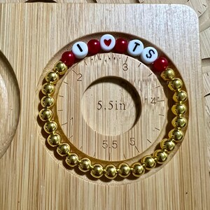 Op de afbeelding: Gouden kralenarmband met de letters "I LOVE TS" in witte en rode kralen. De armband wordt getoond op een houten meetinstrument met een cirkel met een diameter van 14 cm.